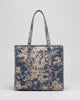 A woven tote bag with a blue and beige floral toile pattern, standing on a gray rug in front of a wooden slat counter inside a shop.