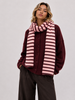A woman wearing a burgundy button-up cardigan, a pink and burgundy striped knit scarf, and dark brown wide-leg trousers, photographed against a neutral light gray studio background.