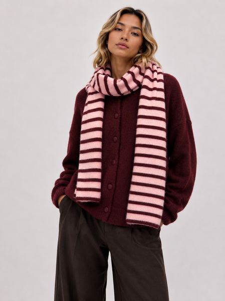A woman wearing a burgundy button-up cardigan, a pink and burgundy striped knit scarf, and dark brown wide-leg trousers, photographed against a neutral light gray studio background.