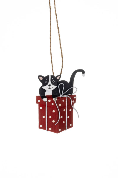A hanging Christmas ornament featuring a black and white cat sitting on top of a red gift box with white polka dots and a white bow, suspended by a natural twine string against a plain white background.