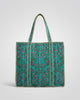 A large woven tote bag with a teal and green floral pattern and gold braided handles, displayed on a gray carpet in front of a wooden slat counter.